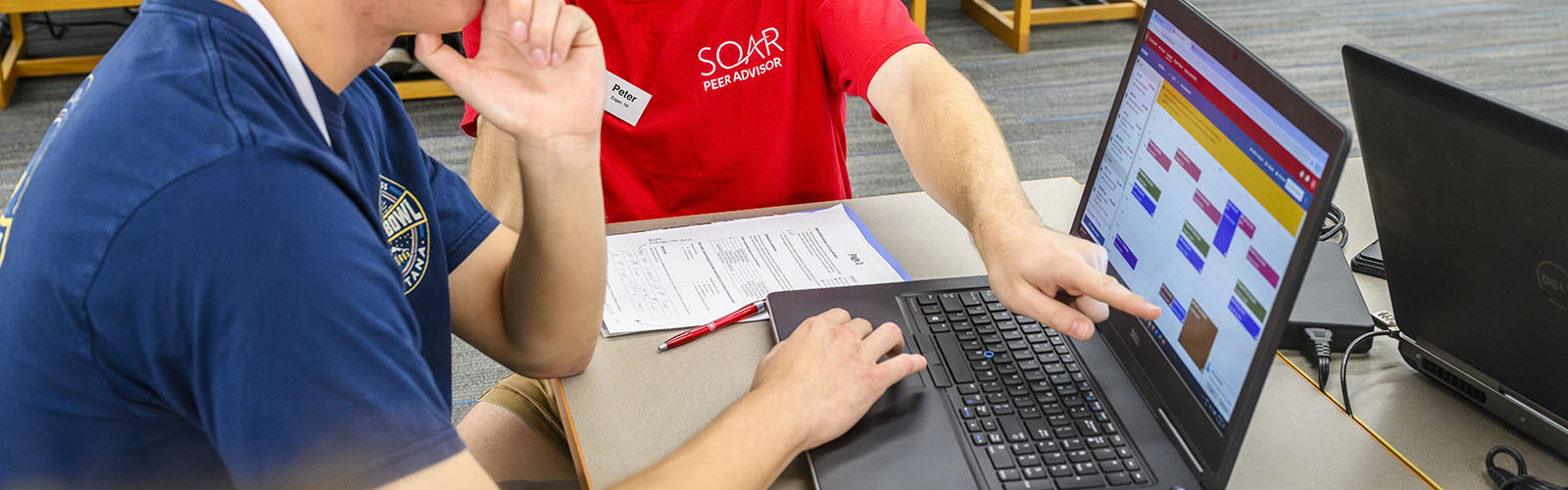On the right side of the image a laptop computer is on a desk while two people look at the screen. One person is pointing to the screen and the other person is using the touchpad on the computer. The computer shows a students schedule.